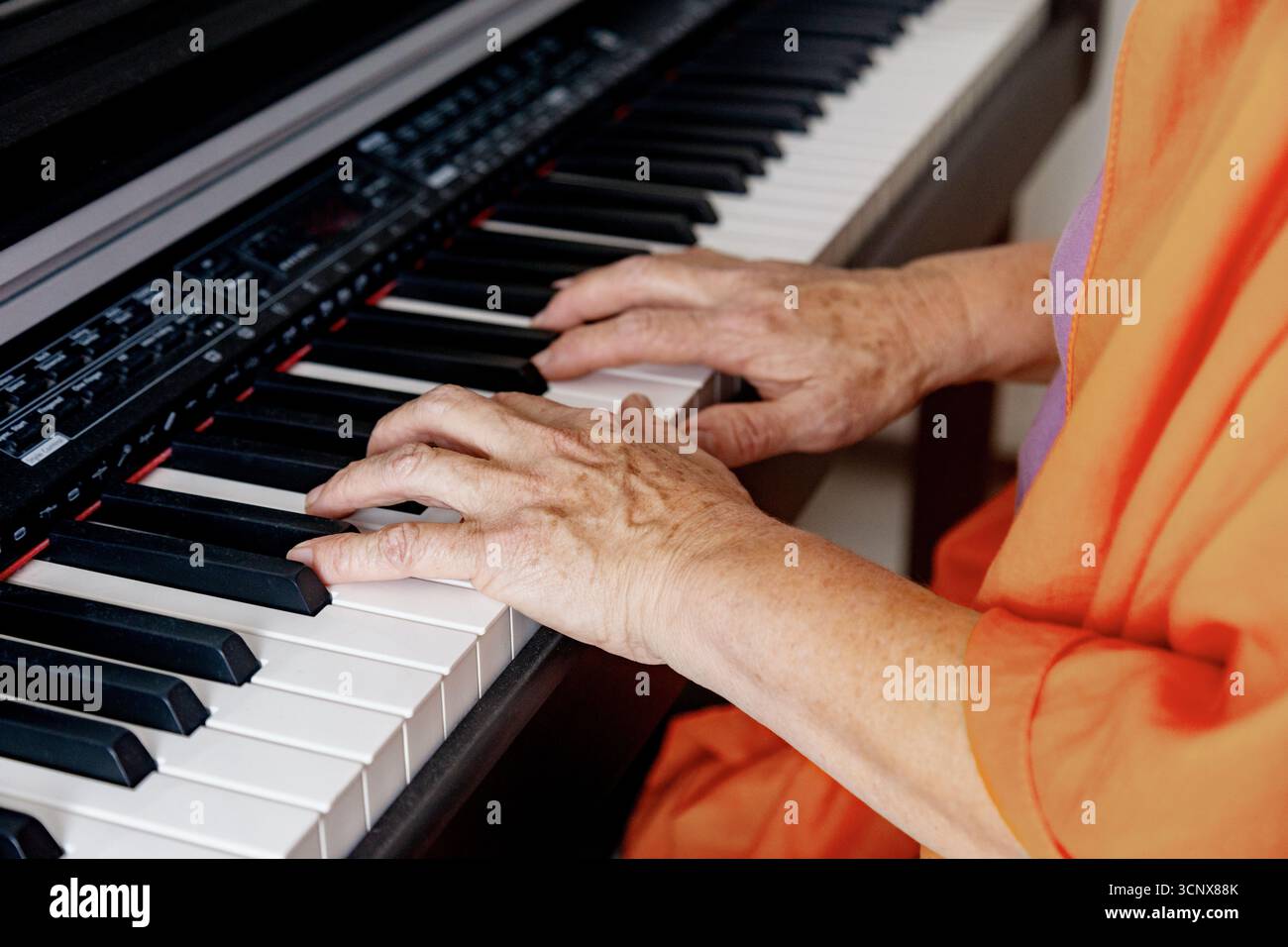 Eine Frau mittleren Alters in orangenem Gewand spielt fröhlich drinnen Klavier und zeigt ihr musikalisches Talent und ihre Leidenschaft für Musik in einem gemütlichen Ambiente Stockfoto