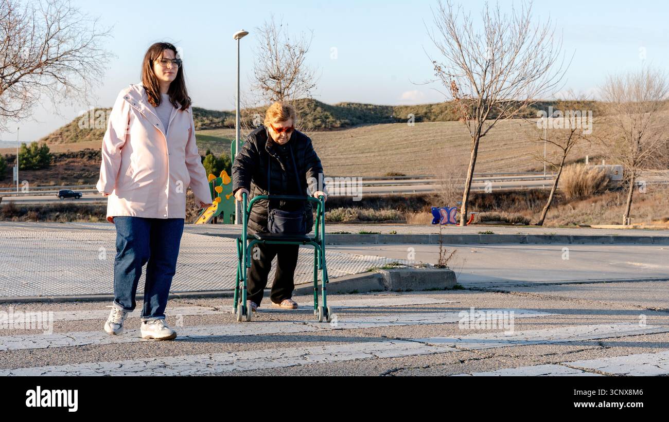 Eine Großmutter benutzt einen Walker, während sie neben ihrer Enkelin über eine ruhige Straße in einer ländlichen Gegend läuft. Die Szene reflektiert Familienunterstützung, Liebe und Stockfoto