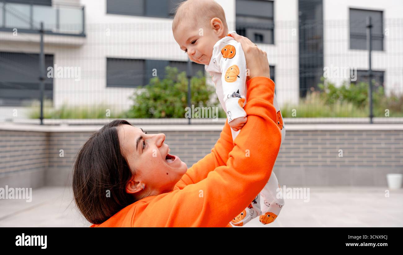 Eine Mutter hält ihren 4 Monate alten Jungen während eines Halloween-Fotoshootings auf einer Terrasse fröhlich hoch. Das Baby trägt einen Kürbis mit dem Thema Stockfoto