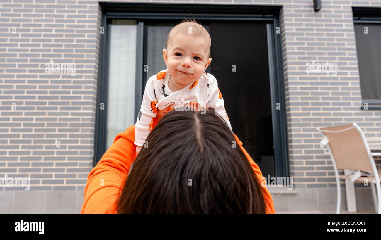 Zugeschnittene Rückansicht einer unerkennbaren Frau, die einen fröhlichen 4 Monate alten Jungen in einem Halloween-Outfit hält, in einer Outdoor-Umgebung lächelnd, s Stockfoto