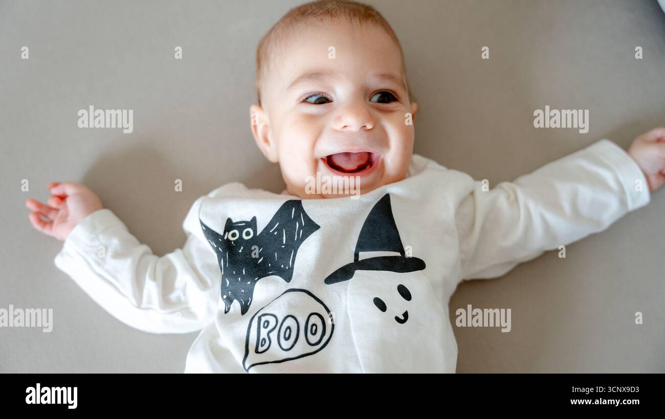 Draufsicht eines 4 Monate alten Jungen liegt auf einem grauen Hintergrund und lächelt in einem Halloween-Outfit mit Fledermaus und Geist mit „BUH“-Text, c Stockfoto