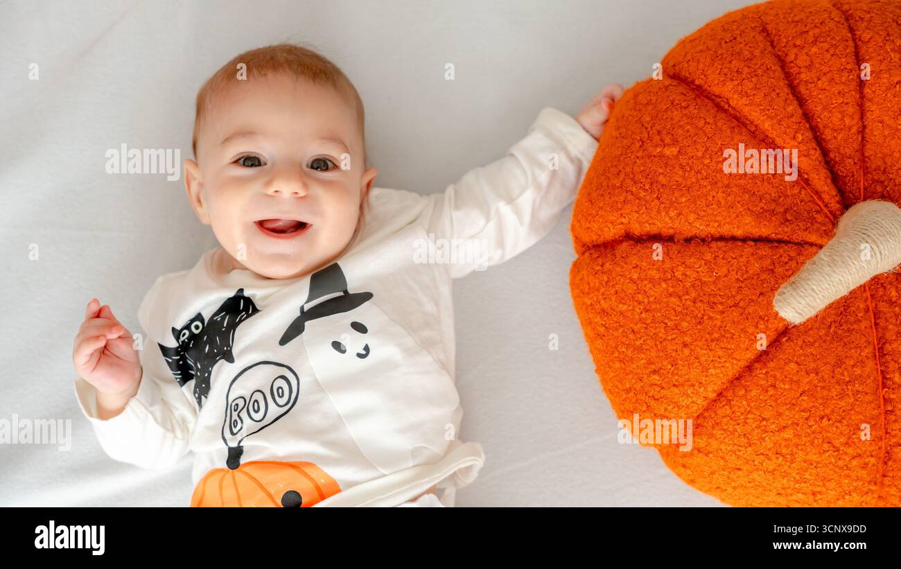 Der Blick von oben auf einen 4 Monate alten Jungen liegt auf weißem Hintergrund, fröhlich lächelnd und trägt ein Halloween-Outfit mit einem Geist und einer Kürbis-Plüschfigur Stockfoto