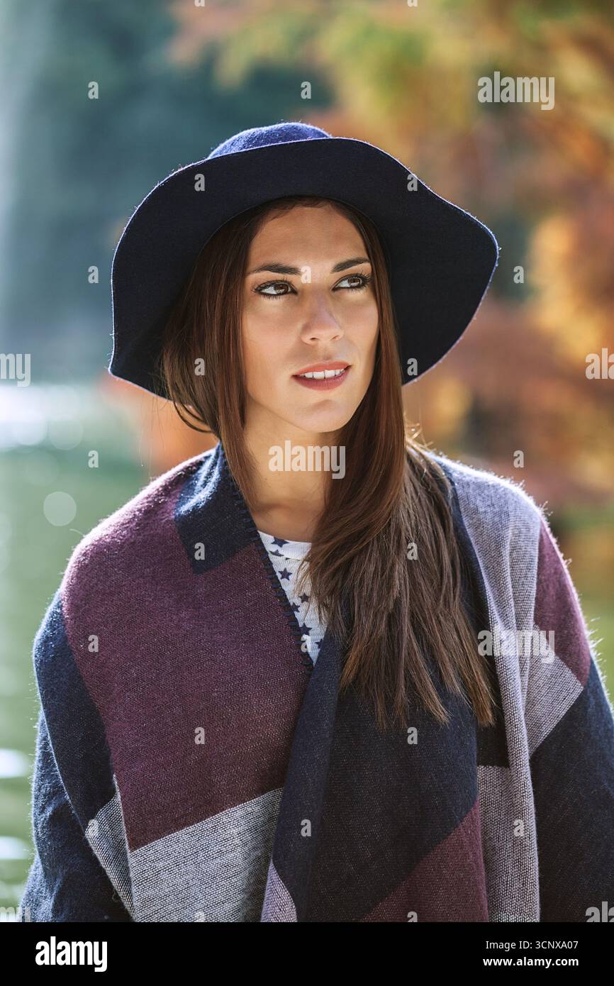 Eine Frau, die einen stylischen Hut mit breiter Krempe und einen gemütlichen Schal trägt, steht draußen und blickt nachdenklich. Der Hintergrund deutet auf eine ruhige, herbstliche Umgebung hin Stockfoto