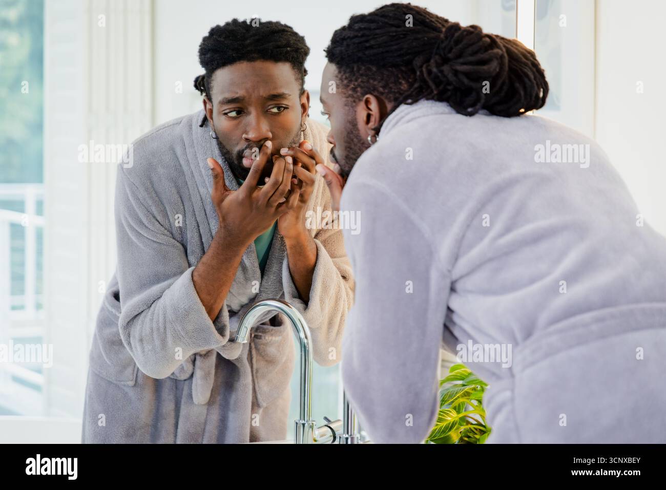 Afroamerikaner beugt sich nach vorne und inspiziert Gesicht im Spiegel am Waschbecken neben Topfpflanzen Stockfoto