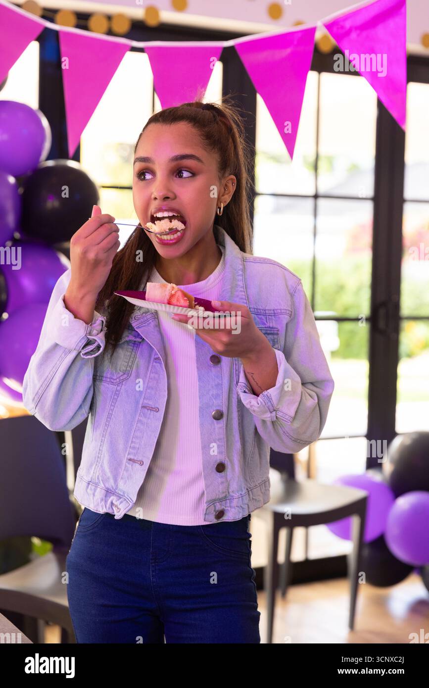 Ein junges Mädchen steht und isst ein Stück Kuchen vom Teller auf einer Party mit Ballons und Bannern Stockfoto