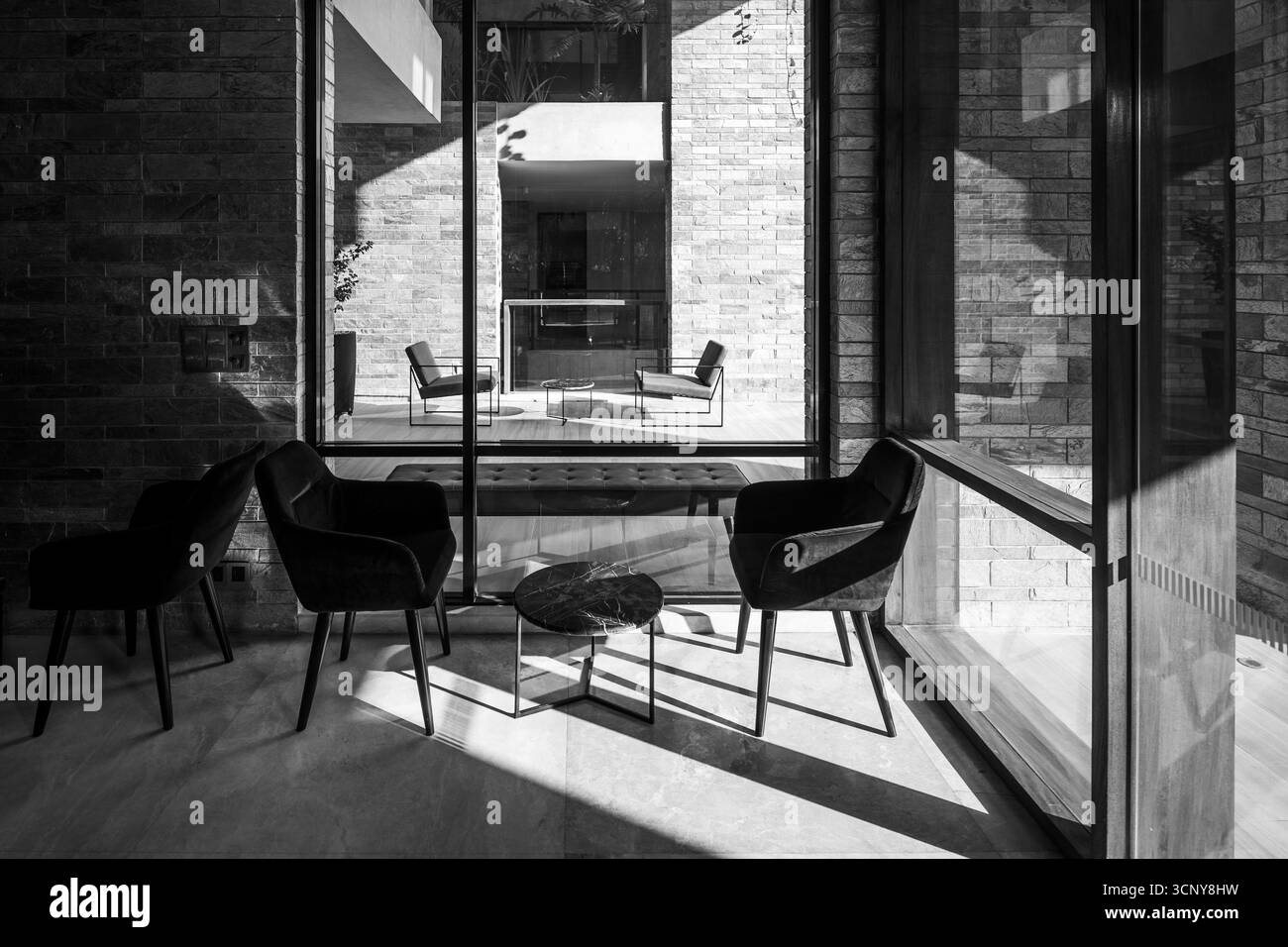Ein eleganter und minimalistischer Indoor-Lounge-Bereich mit weichen Sesseln und einem kleinen Tisch, der durch ein großes Fenster von einer Außenterrasse getrennt ist. Stockfoto