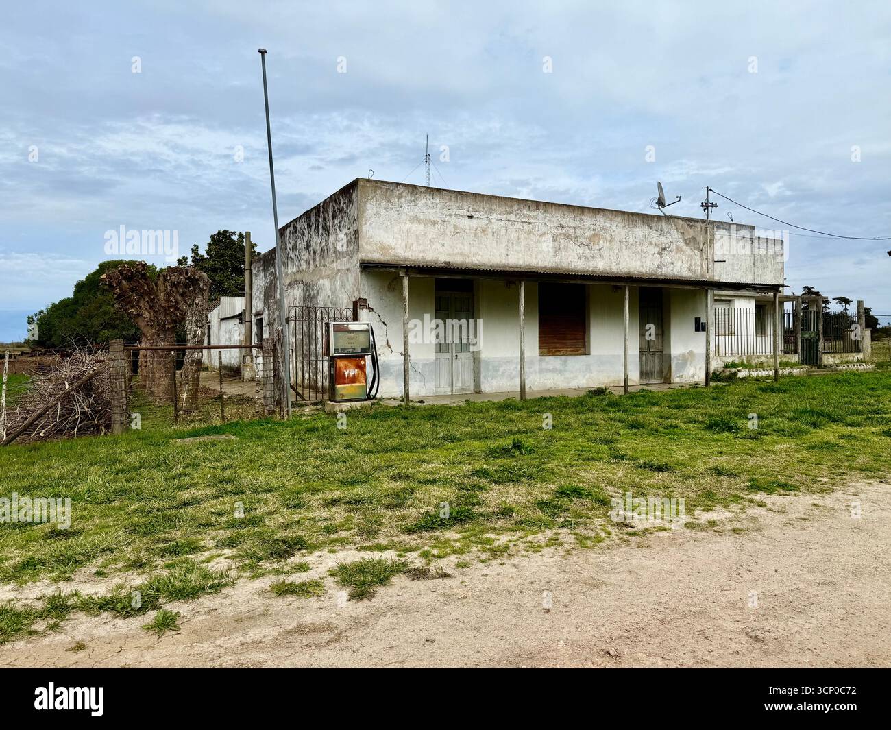 Ländliches Gebäude in Buenos Aires, Argentinien, mit einem rostigen Welldach und abblätternder Farbe, umgeben von einem grasbewachsenen Feld unter bewölktem Himmel. - Smartphone-aufgenommenes Stockfoto