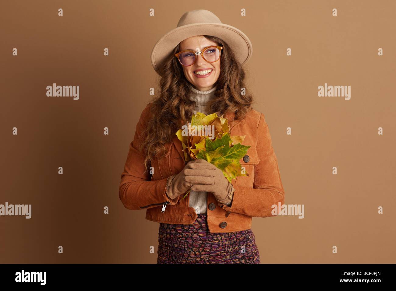 Eine fröhliche Frau in ihren frühen 30ern, gekleidet mit breiter Krempe und Herbstmode, lächelt, während sie leuchtende Herbstblätter vor einem satten braunen bac hält Stockfoto