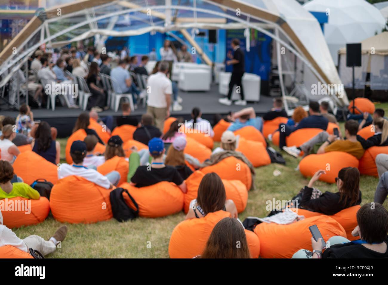 Die Menge entspannt sich im Freien auf orangefarbenen Sitzsäcken, während der Moderator unter Schutz spricht, mit Sitzwänden, lebendiger Veranstaltungsatmosphäre vielfältige Teilnehmer diskutieren und zuhören. Stockfoto
