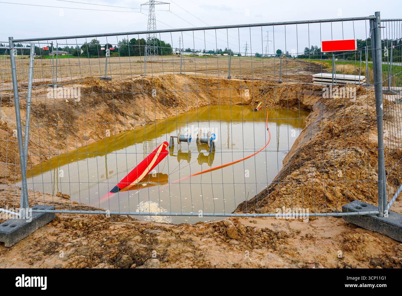 Verfahren zur Verlegung von Stromkabeln in Wellrohren, bei denen hinter einem Sicherheitszaun ein mit Schlammwasser gefüllter Graben mit Spulen, Kabeln und Rohren steht Stockfoto