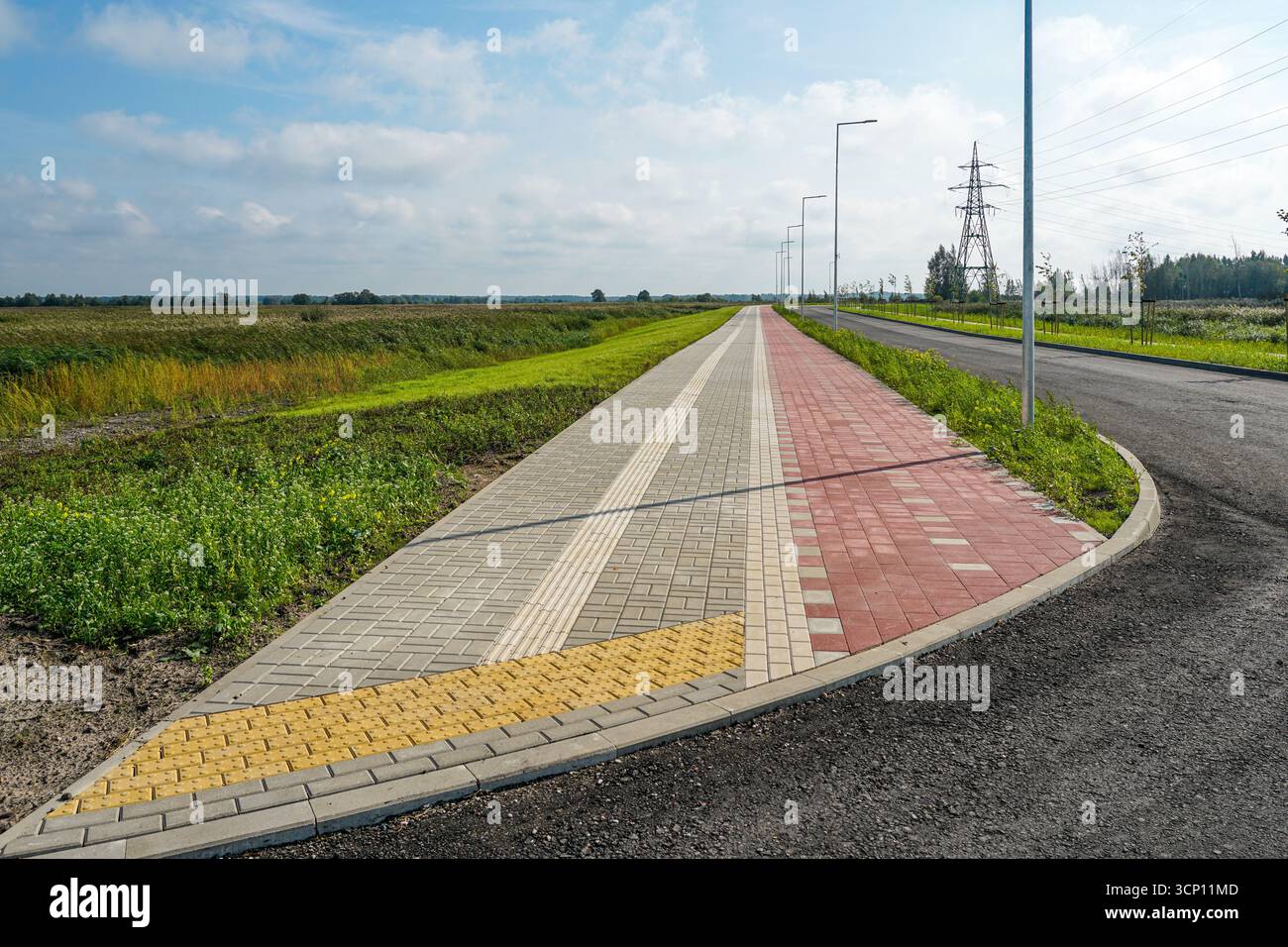 Moderner Fußgänger- und Fahrradweg mit taktilem Pflaster und Straßenlaternen, der entlang einer neuen Straße in ländlicher Landschaft verläuft Stockfoto