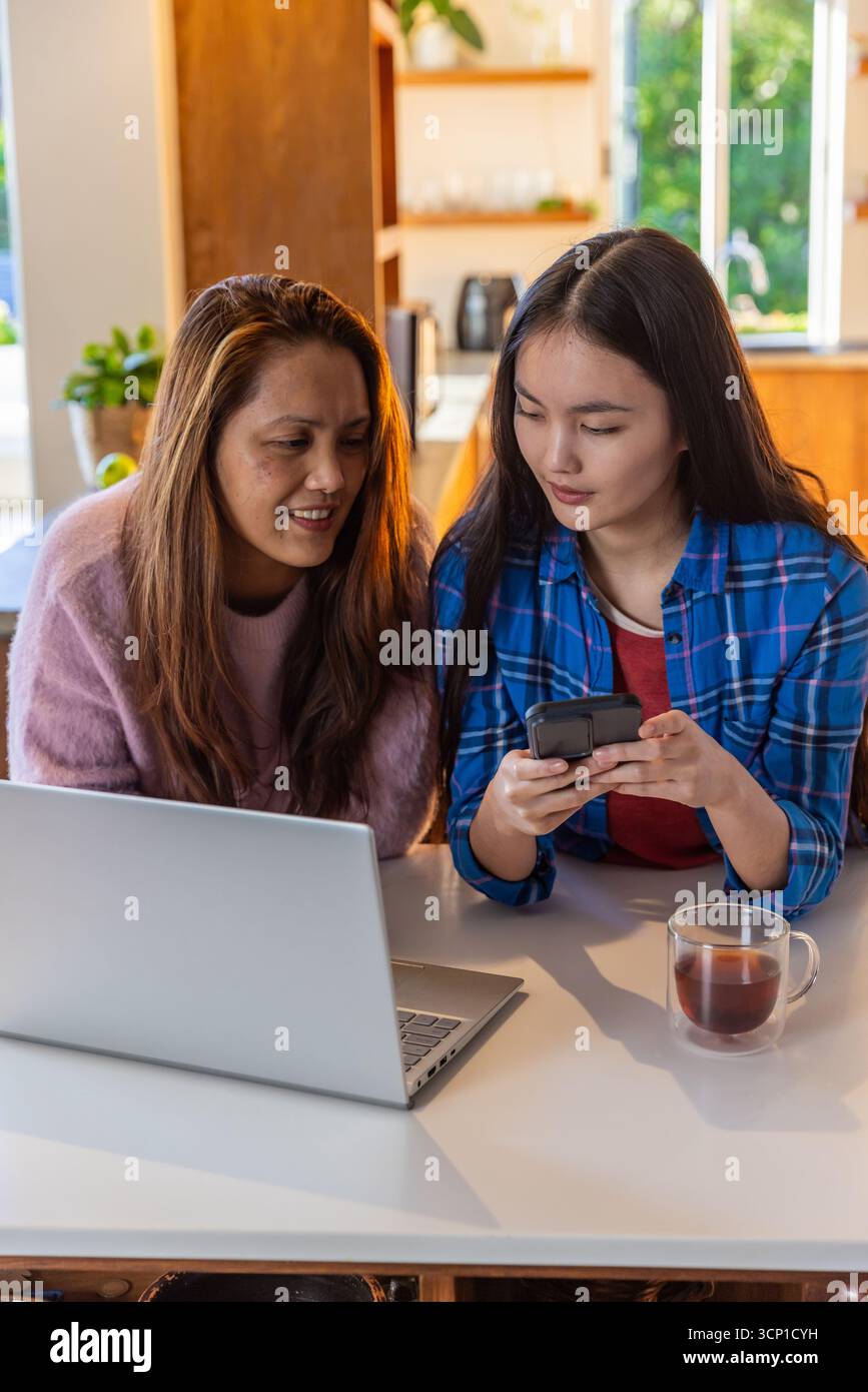 Asiatische Mutter und Tochter teilen sich ihr Smartphone, während sie am Tisch am Laptop arbeiten Stockfoto