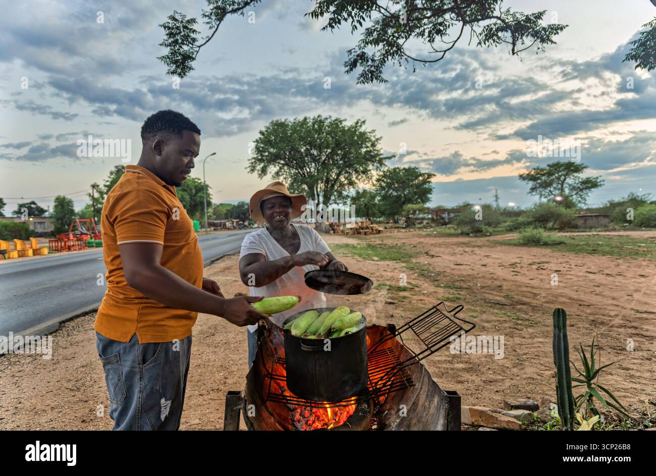 Afrikanischer Straßenverkäufer, Kunde, der gekochten Mais kauft, Frau, die Mais auf dem Feuer in einem Topf auf dem Grill am Straßenrand im Dorf verkauft Stockfoto