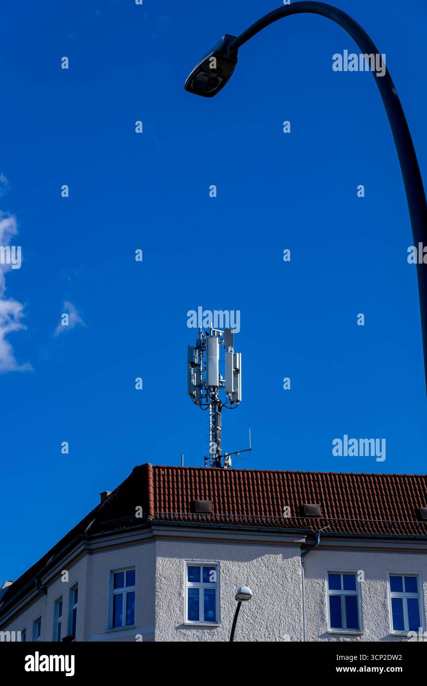 Vertikales Bild der 5G-Mobilfunkantenne (5G-Mast) auf dem städtischen Dach in Berlin mit gekrümmter Straßenlaterne im Vordergrund, klarem blauem Hintergrund. Stockfoto
