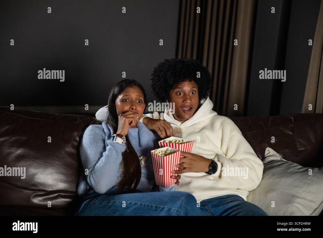 Verschiedene Paare sehen sich intensive Filme an, während sie gestreifte Popcorn-Wannen auf einem Ledersofa halten Stockfoto