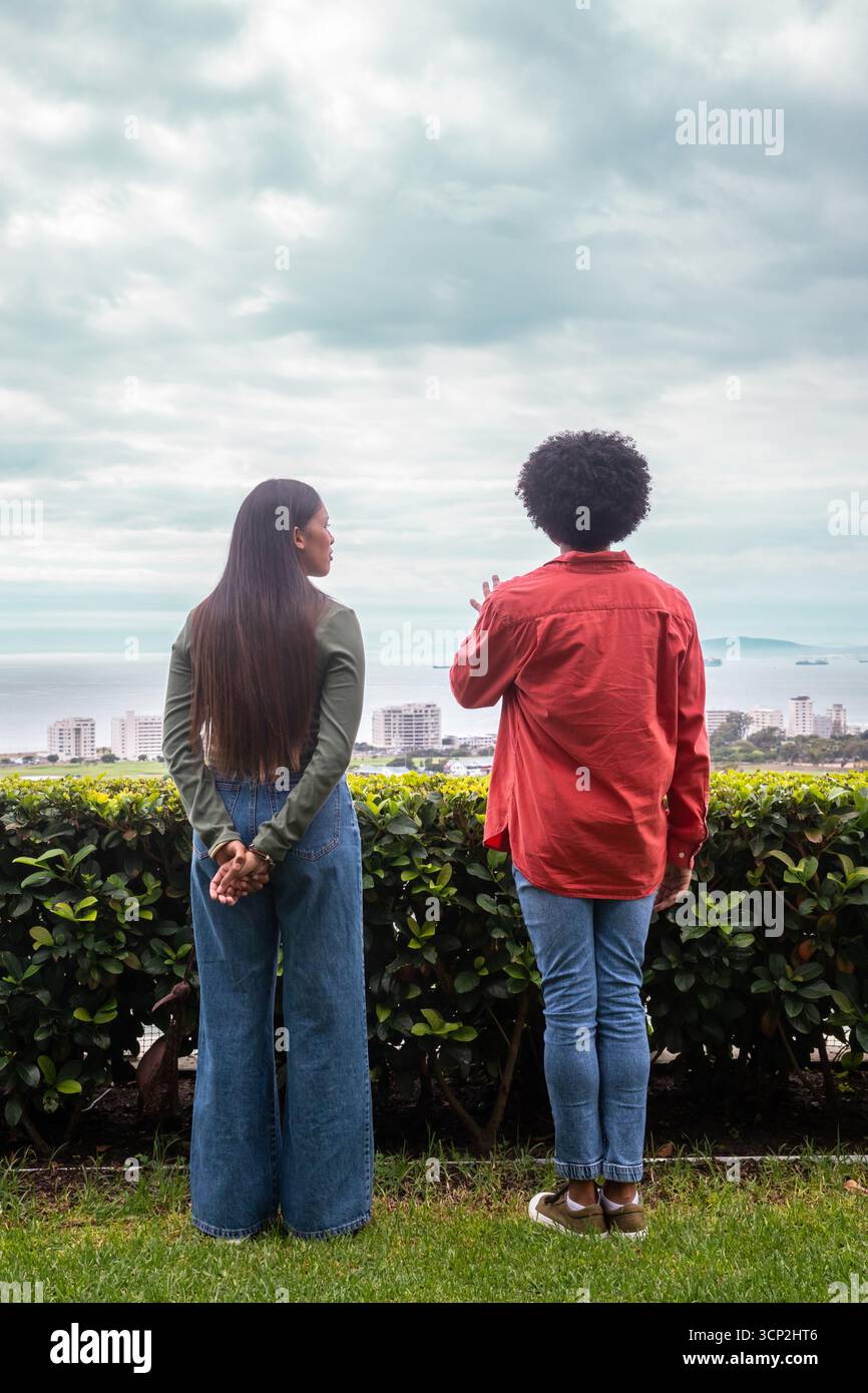 Verschiedene Freunde stehen auf einer grasbewachsenen Terrasse mit Blick auf die Stadt, das Meer, mit Hecke unter bewölktem Himmel Stockfoto