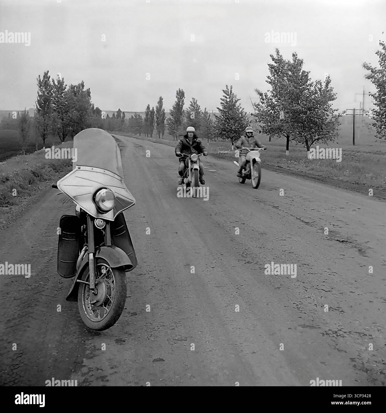 Zwei sowjetische Motorradfahrer auf Enduro-Fahrrädern werden 1980 während eines Rallyefahrens in der Nähe von Sloviansk, der ukrainischen SSR, auf einer Landstraße gesehen. Das Foto wurde aus der Perspektive eines dritten, schwereren Straßenmotorrads mit großer Windschutzscheibe aufgenommen, wahrscheinlich einer Begleitperson oder eines offiziellen Fahrrads, das auf der Schulter geparkt ist. Dieses stimmungsvolle Schwarz-weiß-Bild fängt den Geist des Abenteuers und das offene Straßengefühl eines klassischen Motorsport-Events in der UdSSR ein Stockfoto
