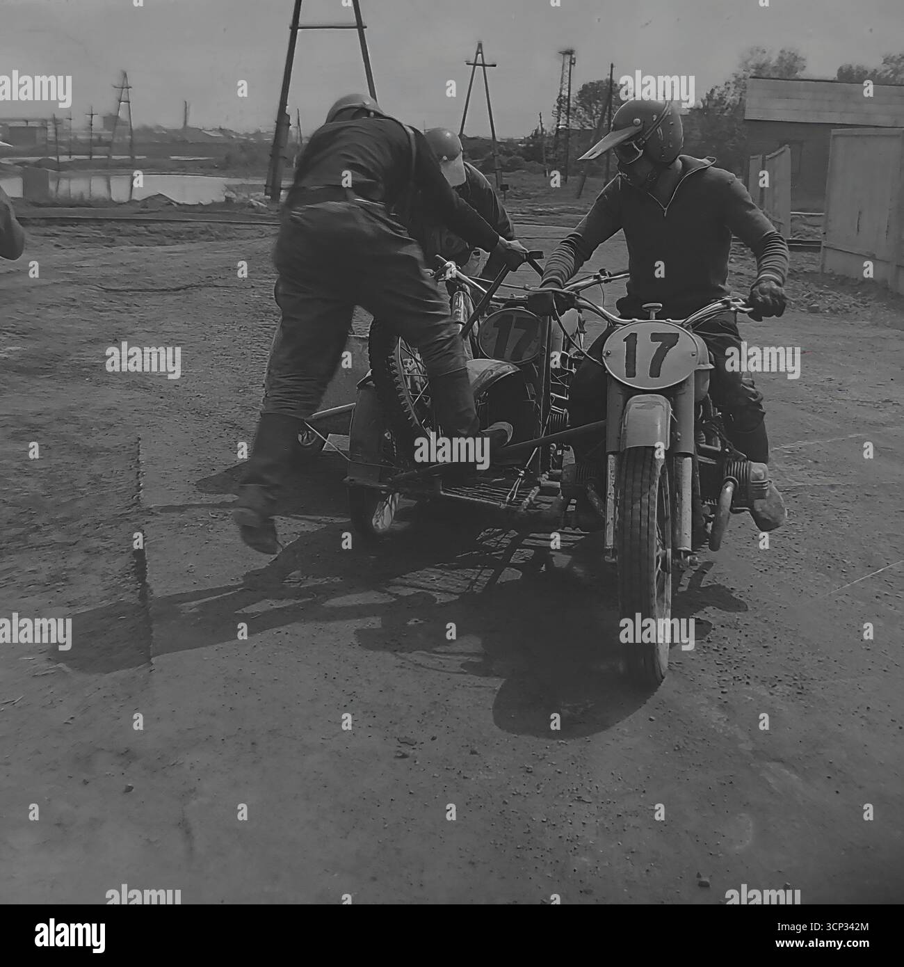 Ein aufregender Action-Schuss aus dem Jahr 1980 fängt ein sowjetisches Motocross-Team mit der Nummer 17 ein, das in Sloviansk, der ukrainischen SSR, an einem Rennen teilnimmt. Der Beifahrer, auch „Affe“ genannt, wird sportlich gesehen, wie er sein Gewicht verlagert und dabei hilft, die Maschine zu schieben, was die unglaubliche Teamarbeit und die körperlichen Anforderungen dieses Vintage-Motorsports demonstriert. Dieses dynamische Schwarzweiß-Foto friert einen Moment intensiver Anstrengung und Koordination auf der Feldbahn ein Stockfoto