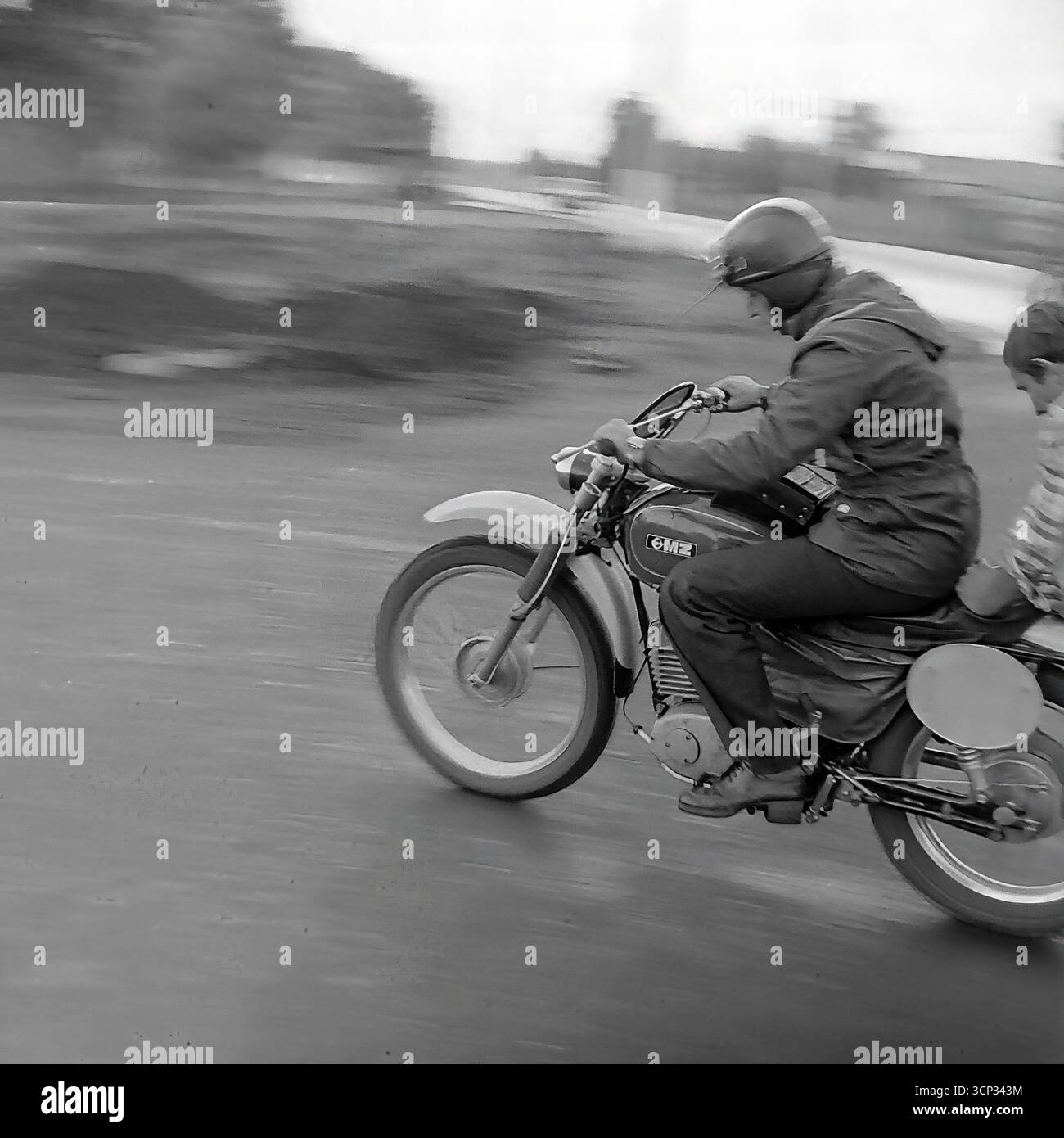 Ein atemberaubender Schwenkschuss aus dem Jahr 1980 fängt einen sowjetischen Rennfahrer auf einem klassischen ČZ-Motorrad ein, der einem Passagier bei einem Motorsportevent in Sloviansk, der ukrainischen SSR, eine Fahrt zum Mitfahrer gibt. Fahrer und Beifahrer sind im scharfen Fokus vor einem wunderschönen bewegungsunscharfen Hintergrund gefroren, der perfekt die aufregende Geschwindigkeit und einen einzigartigen Moment des gemeinsamen Erlebnisses vermittelt. Dieses dynamische Schwarzweiß-Foto ist ein Beweis für die Begeisterung und den Gemeinschaftsgeist eines klassischen Rennsports aus der Sowjetzeit Stockfoto