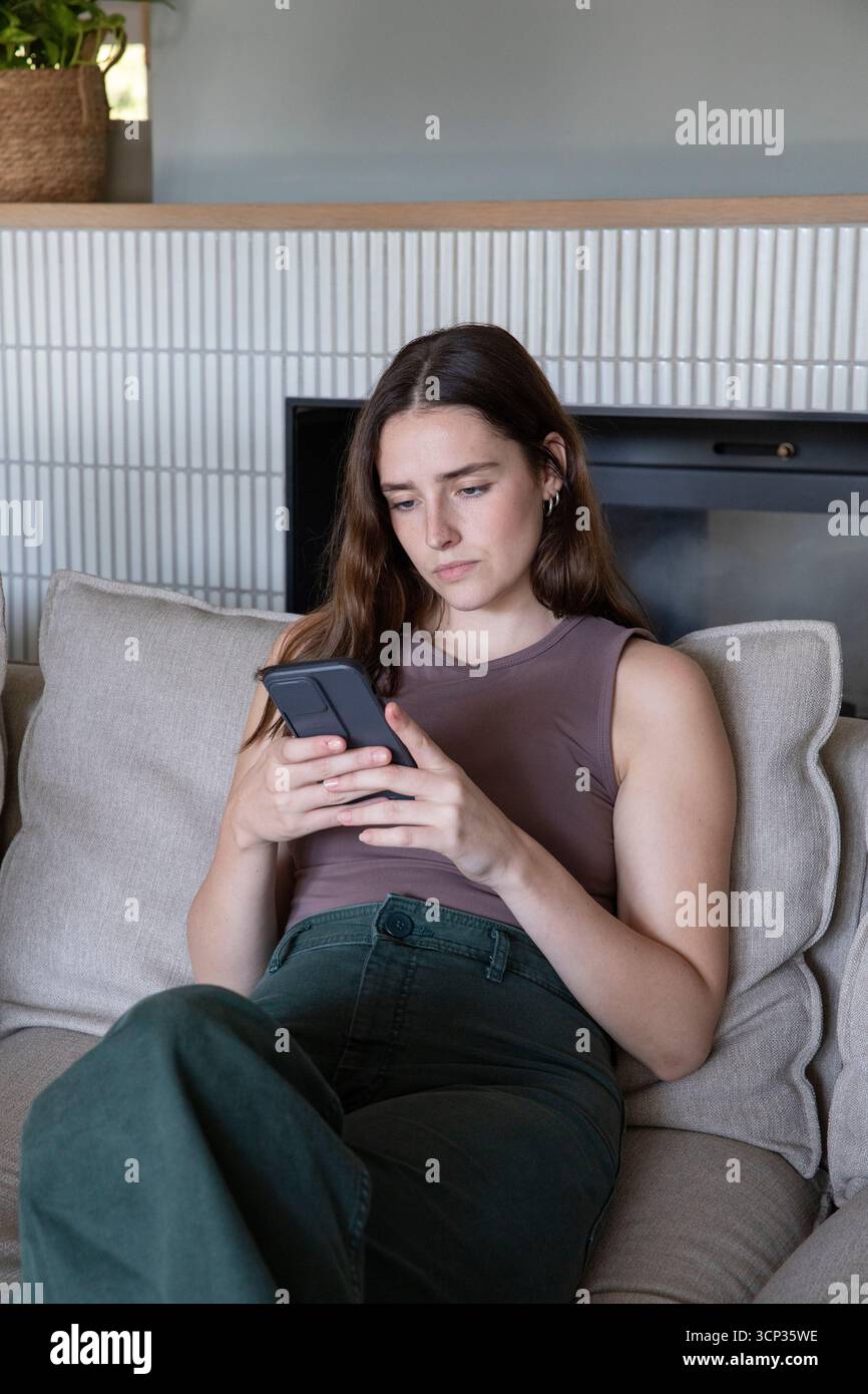 Eine Frau in ihren Zwanzigern hält ein Smartphone und lehnt sich auf einem beigefarbenen Sofa am Kamin im Wohnzimmer Stockfoto