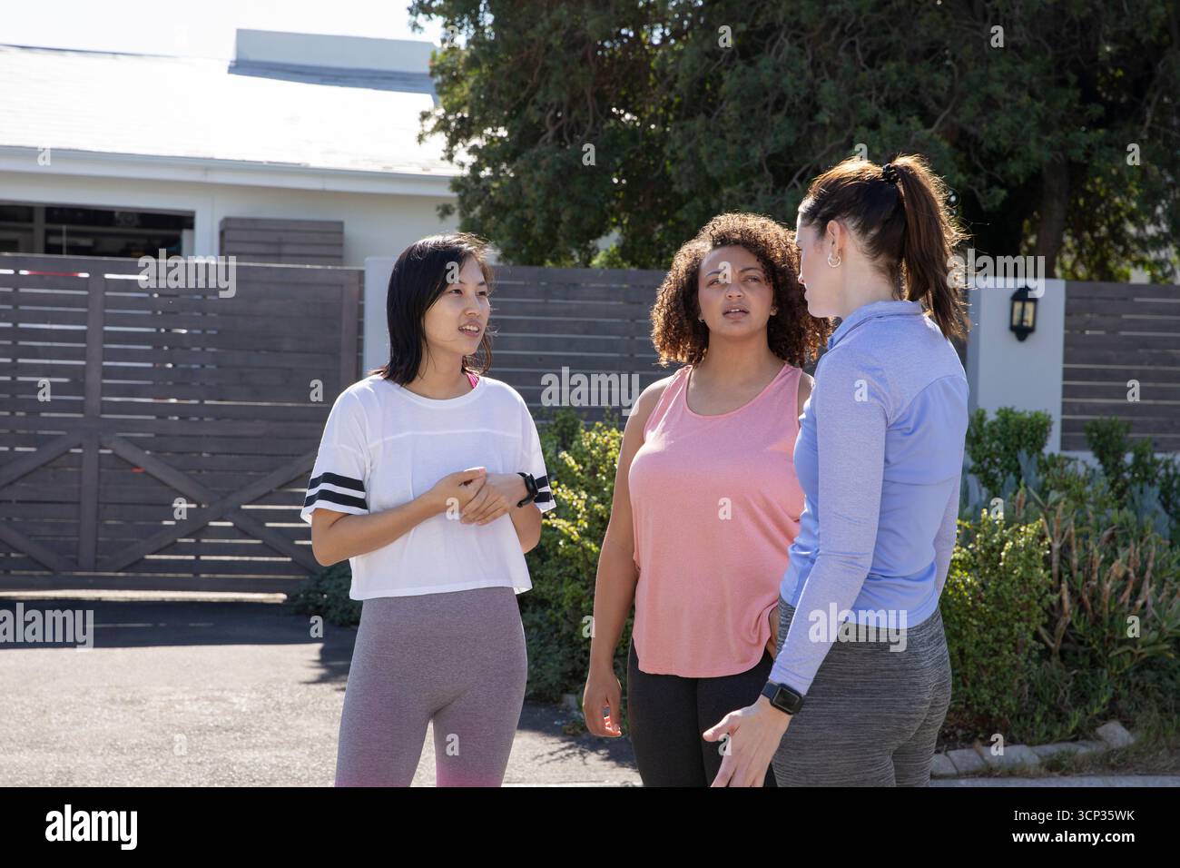 Verschiedene Freundinnen unterhalten sich, während sie auf einer gepflasterten Einfahrt am grauen Tor mit Smartwatches stehen Stockfoto