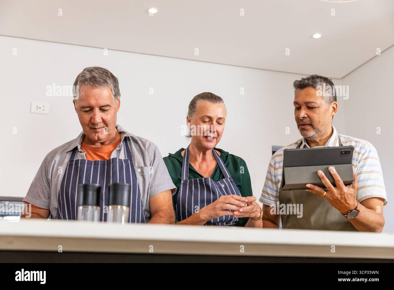Verschiedene ältere Freunde kochen auf der weißen Kücheninsel mit Schürzen, die Rezepte auf einem Tablet lesen Stockfoto