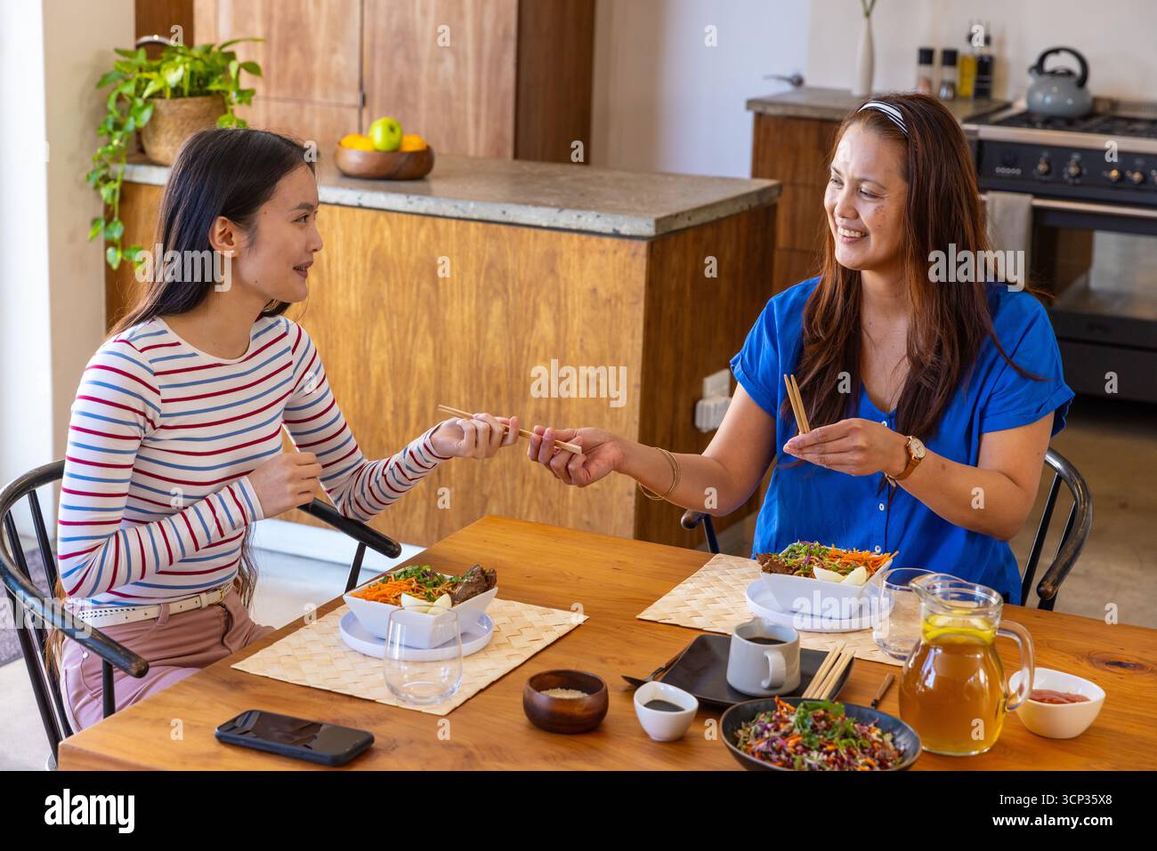 Asiatische Mutter und Tochter tauschen Essstäbchen über Salatschalen am Esstisch mit Eistee aus Stockfoto