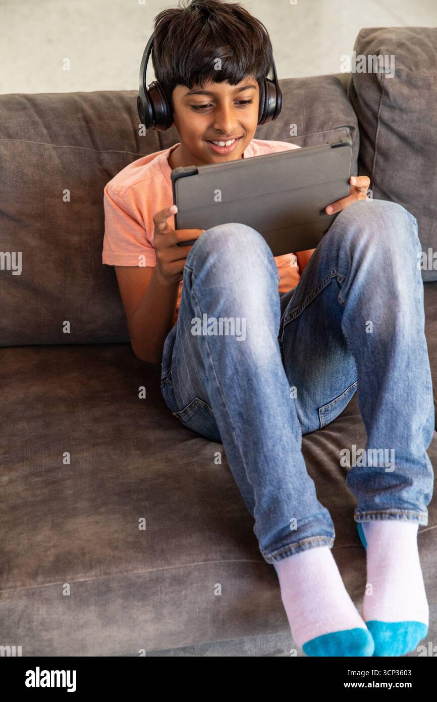 Indischer Junge, der zu Hause auf dem Sofa sitzt und ein Tablet mit Kopfhörern in der Nähe der Kissen verwendet Stockfoto