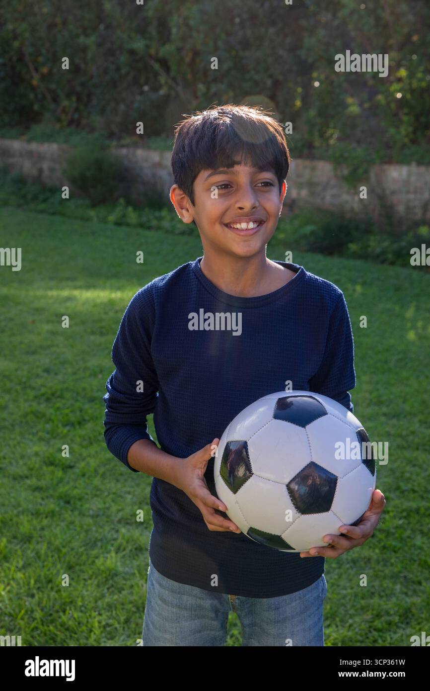 Asiatischer Junge im Schulalter, der Fußball auf dem Garten mit Steinmauerhecken hält Stockfoto