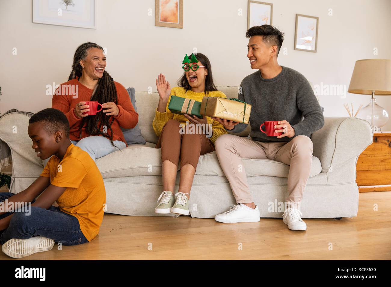 Eine vielfältige Familie, die zu Hause auf der Couch sitzt, lacht und Tassen und Geschenke hält, Junge spielt Stockfoto