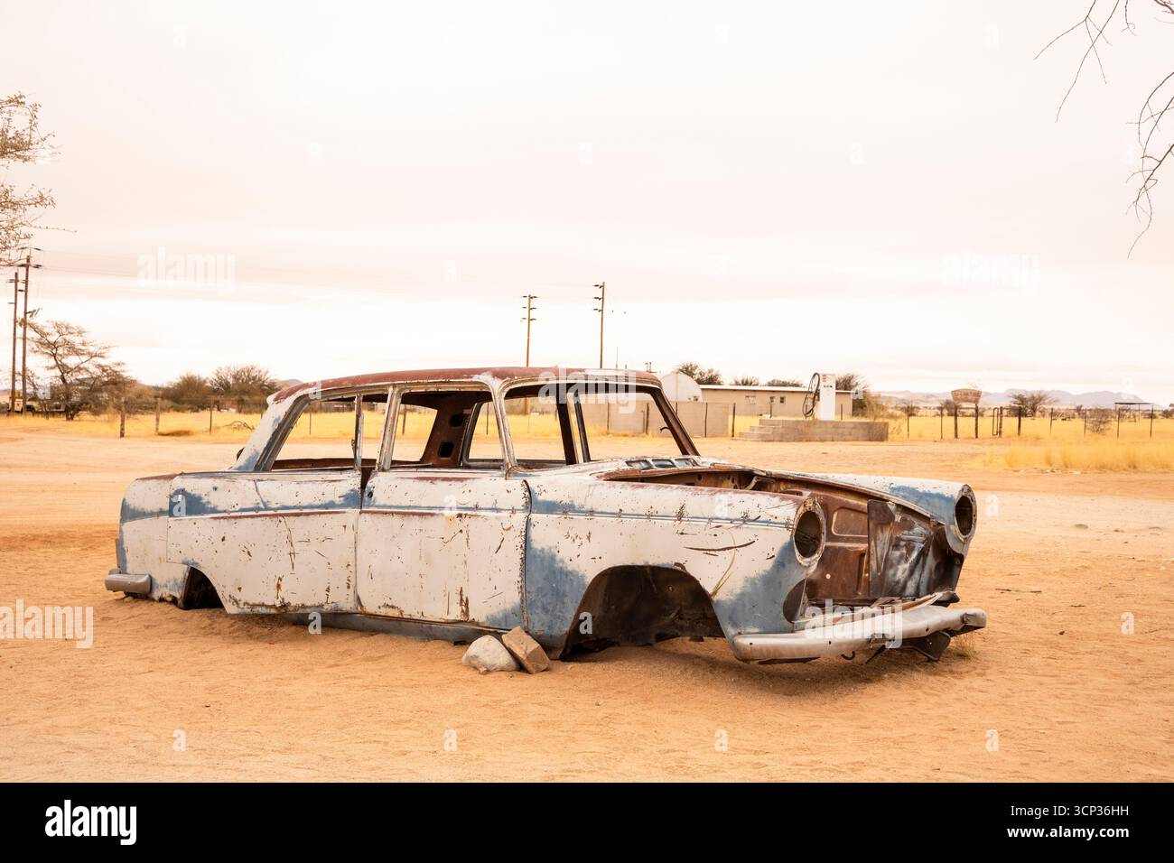 Auf dem Autofriedhof von Solitaire in Namibia befinden sich rostige Oldtimer in der Nähe kleiner Wüstengebäude, die vor Dünen und Bergen liegen und den Charme der Wüste einfangen Stockfoto