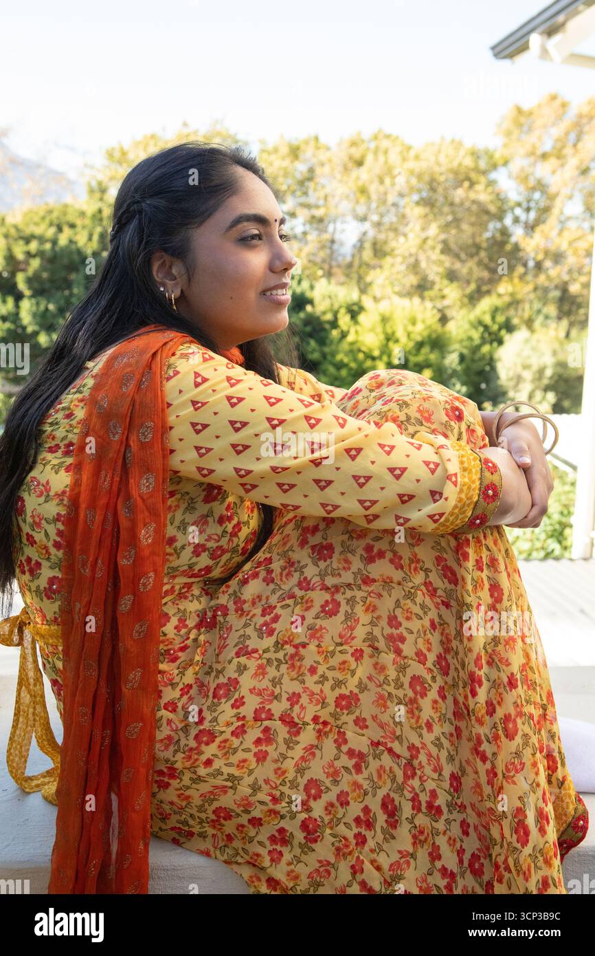 Indische Frau umarmt Knie und blickt auf die Veranda Geländer in Salwar-Kameez mit Reifen und Armreifen Stockfoto