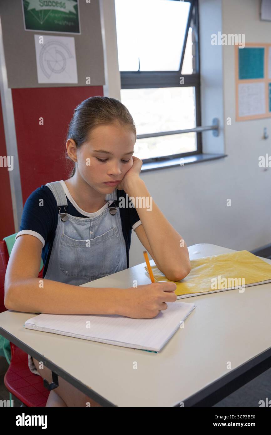 Ein junges Mädchen, das Overalls trägt und in Notizbuch mit Bleistift schreibt, am Schulpult neben dem Fenstergeländer Stockfoto