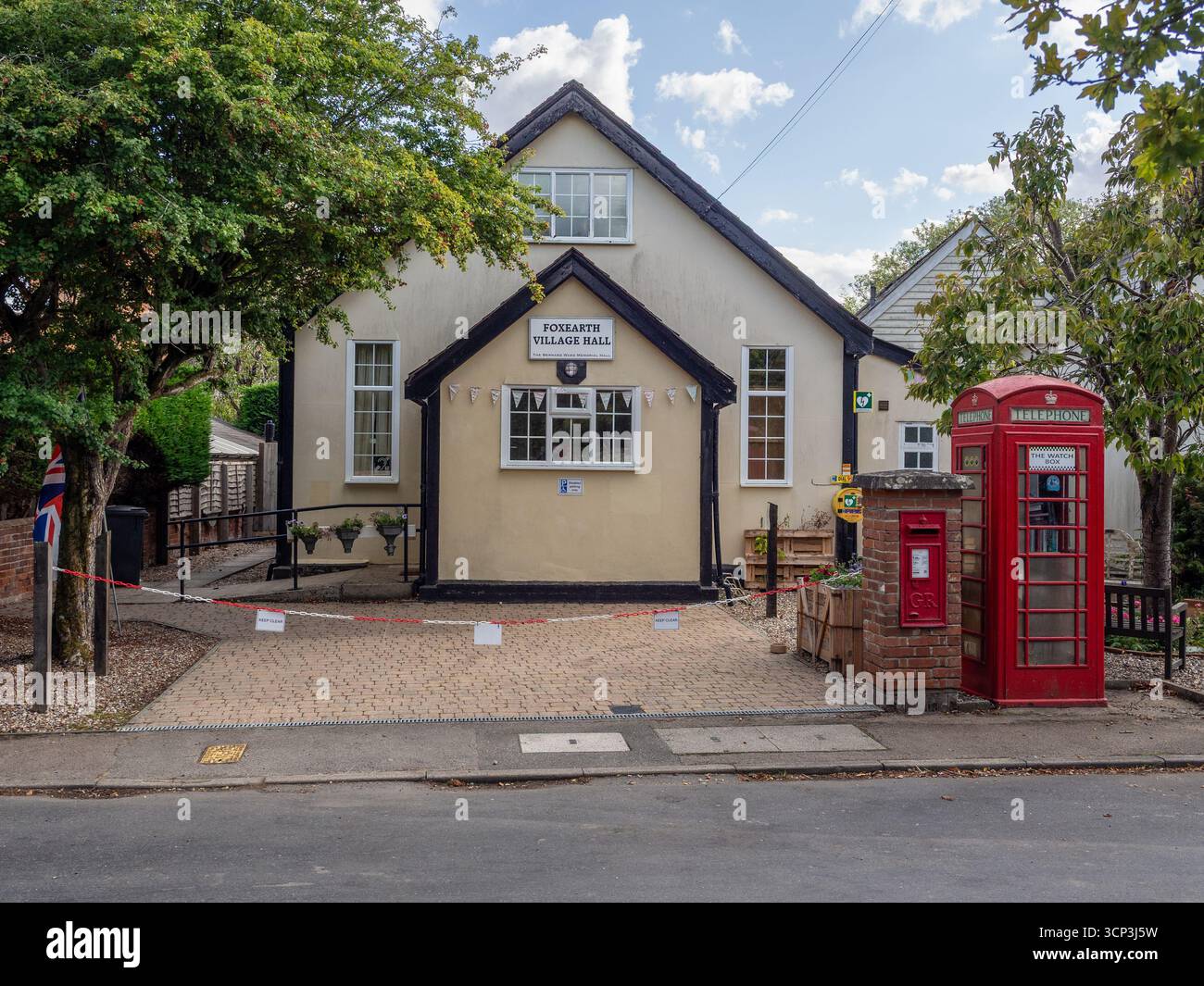 FoxEarth Village Hall in Essex, Großbritannien, mit einem roten Briefkasten der Royal Mail und einem umfunktionierten Telefonfach, das heute als Buchaustausch genutzt wird. Stockfoto