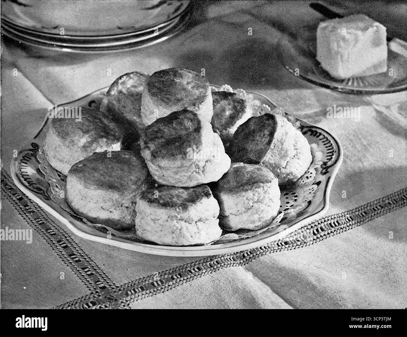 Nahaufnahme eines Gerichts mit Scones (in einem vulkanischen Ofen gekocht) auf einem Esstisch und eines weiteren mit Butter. Aus einem gedruckten Foto, das ursprünglich in der Publikation erschien, The Vulcan Cook Book, das 1936 von der Vulcan Stove Company in Exeter, England, veröffentlicht wurde. Stockfoto