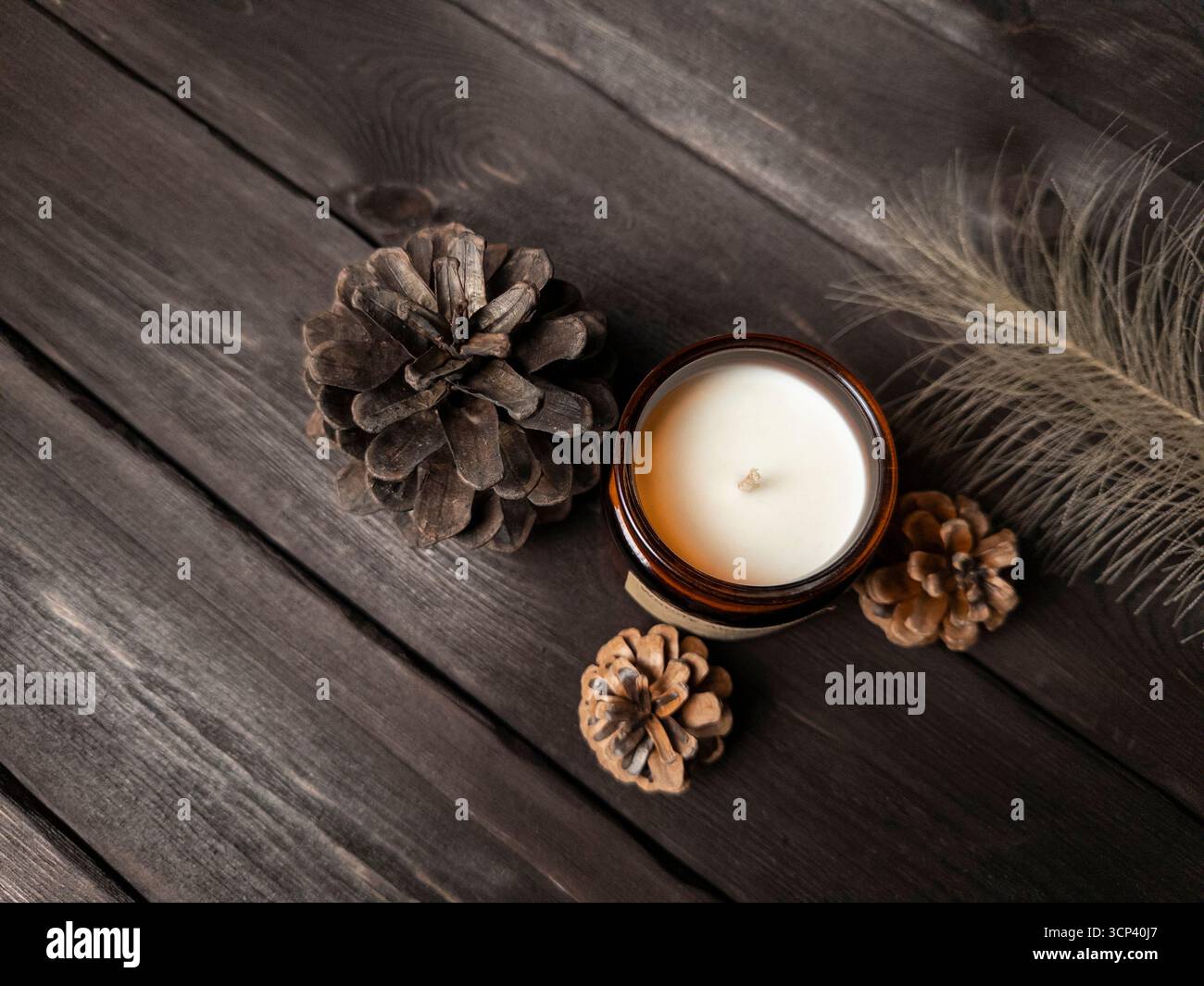 Ein minimalistischer Flachbau mit brauner Glaskerze, Tannenzapfen und einer Feder auf dunkler Holzoberfläche. Ideal für saisonale, Wellness- oder Innenräume Stockfoto