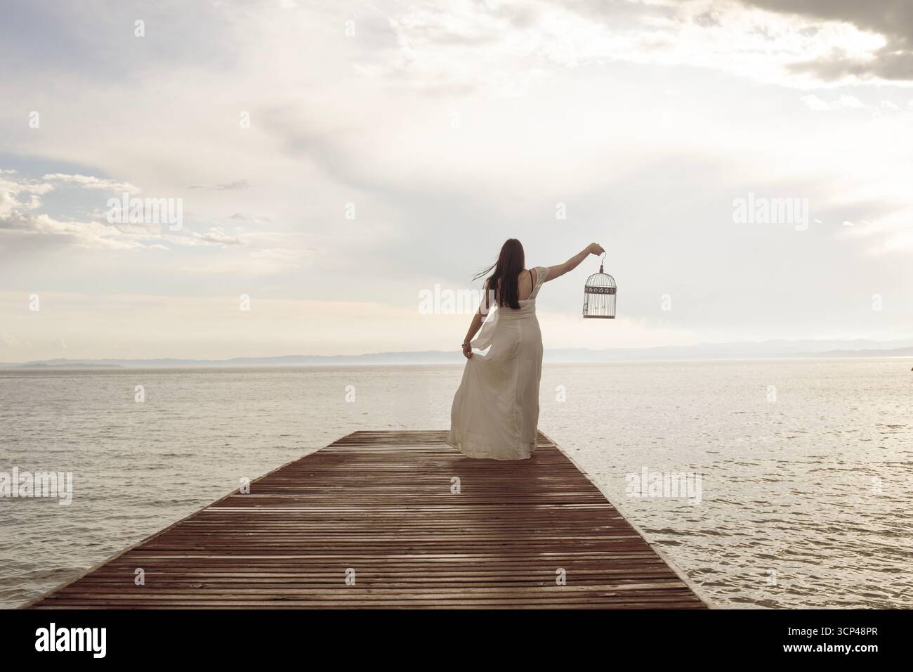 Elegante Frau in einem langen weißen Kleid geht mit einem leeren Käfig zum Meer. Stockfoto