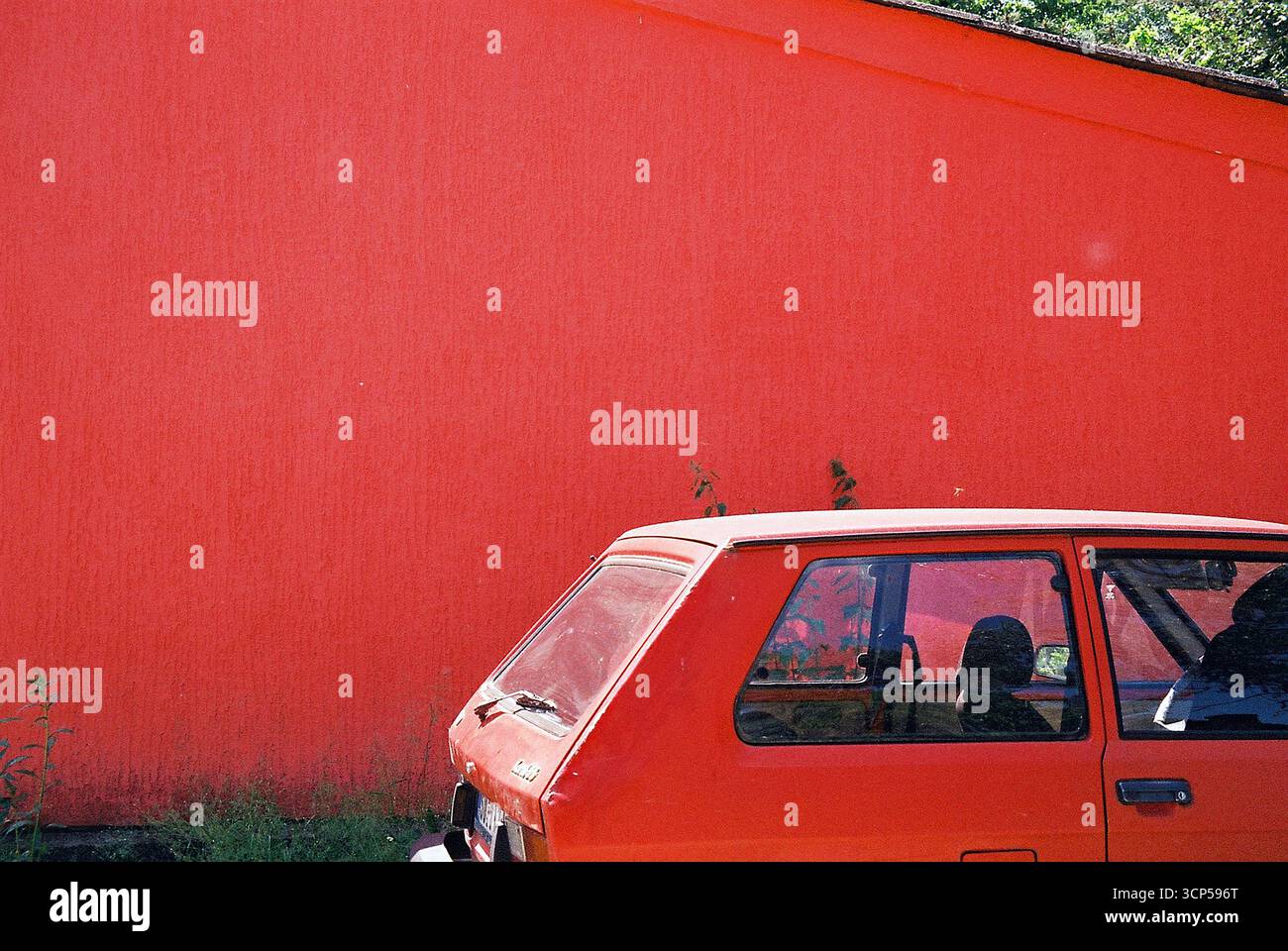 Rotes Yugo-Auto parkte vor einer roten Mauer und roten Wochenendhäusern. Analoges Foto mit auffälliger Farbharmonie und nostalgischer Atmosphäre. Stockfoto