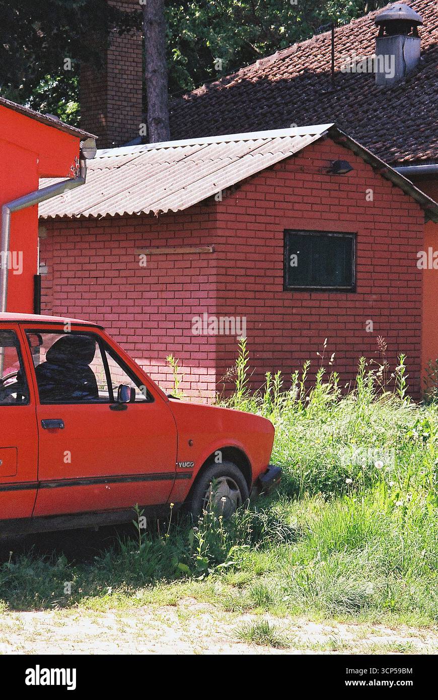 Rotes Yugo-Auto parkte vor einer roten Mauer und roten Wochenendhäusern. Analoges Foto mit auffälliger Farbharmonie und nostalgischer Atmosphäre. Stockfoto
