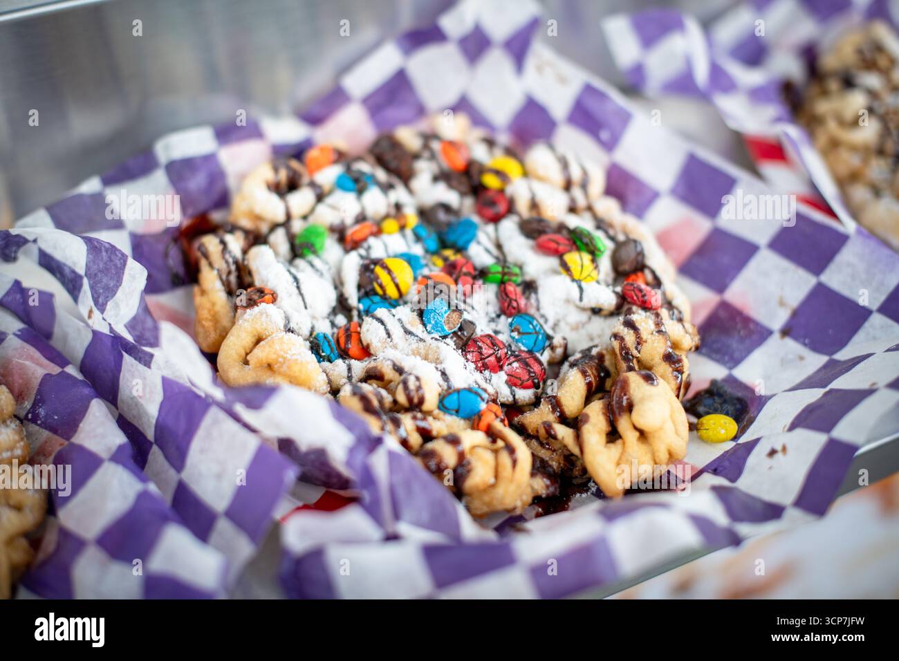 Trichterkuchen mit M&Ms-Schokoladendrizzle Frederick Fairgrounds Frederick | FREDERICK, Md. — Ein mit Puderzucker, M&M's und Schokoladendrizzle überzogener Trichterkuchen liegt in einem lila karierten Papptablett auf den Frederick Fairgrounds, dem Zuhause des Great Frederick Fair. Trichterkuchen, die durch das Gießen von Teig durch einen Trichter in heißes Öl hergestellt werden, sind ein fester Bestandteil des Festessens auf amerikanischen Jahrmärkten und Landwirtschaftsmessen und gehen auf die kulinarischen Traditionen der Pennsylvania Dutch zurück. Stockfoto