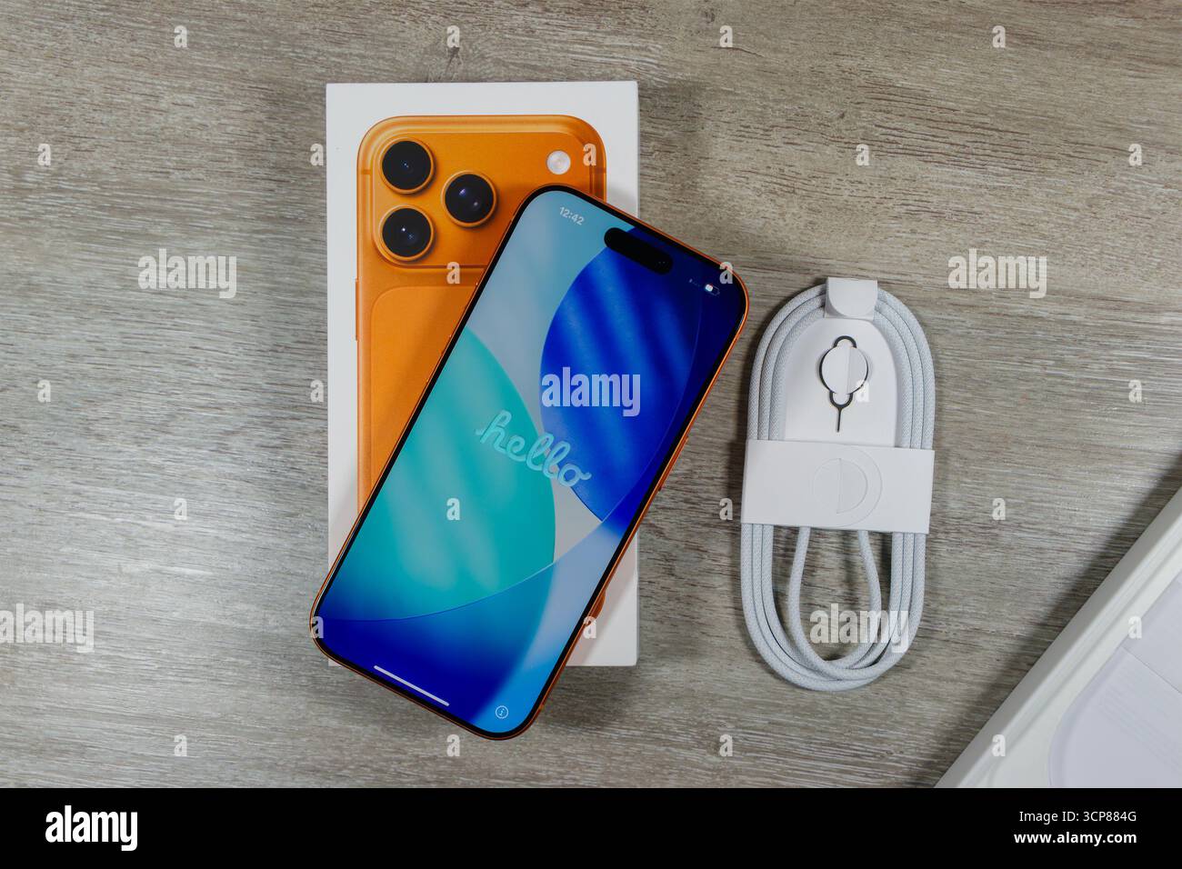 Das neue iPhone 17 Pro Smartphone mit orangefarbener Rückseite und blauem Bildschirm, auf dem „Hallo“ angezeigt wird, befindet sich auf der Box mit dem Ladekabel auf einer Holzoberfläche Stockfoto