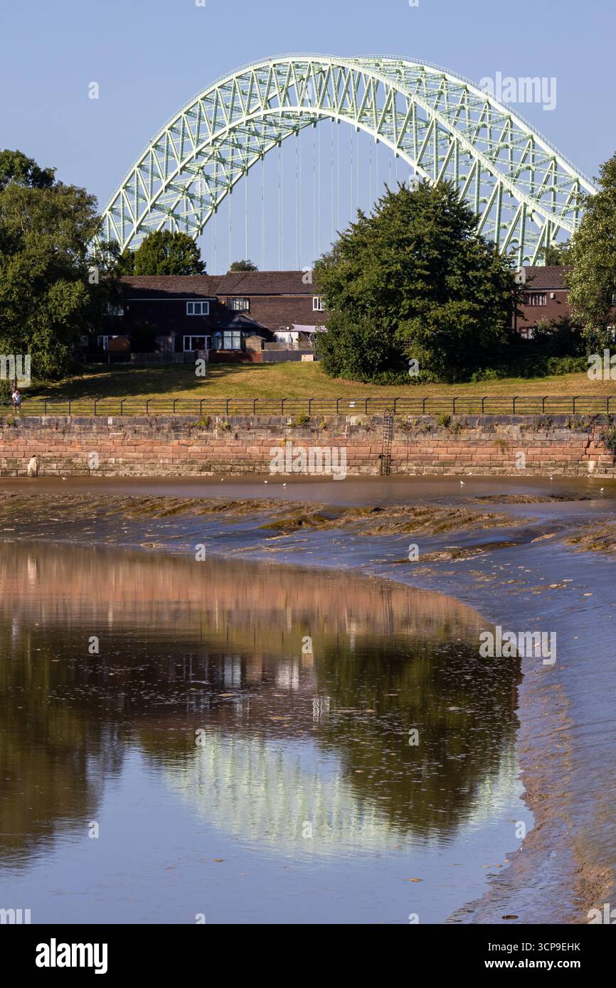 Blick auf den Bogen der Silver Jubilee Bridge bei Runcorn, mit nahe gelegenen Häusern und Bäumen am Flussufer, die sich bei Ebbe im Wasser spiegeln Stockfoto