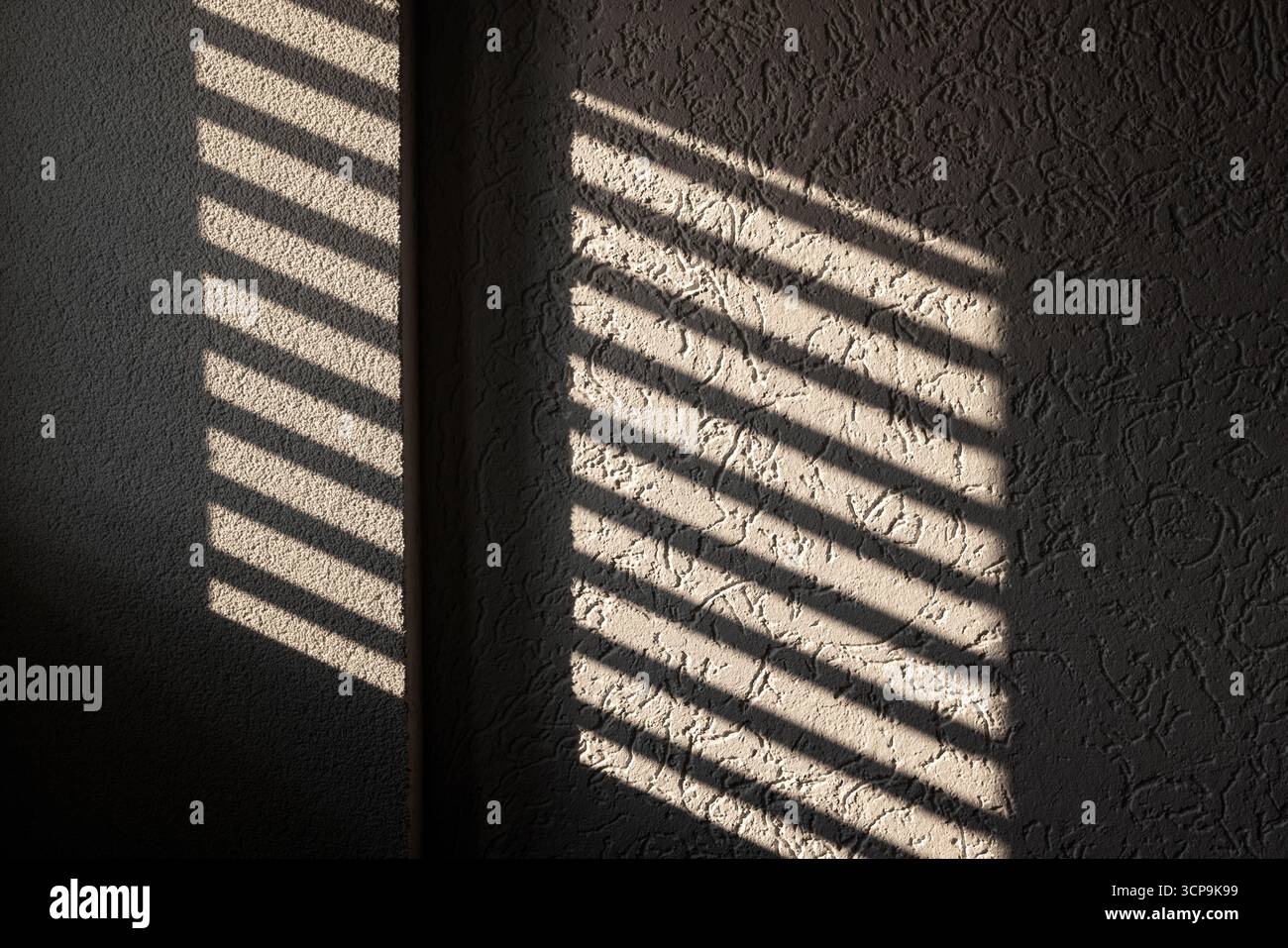 Eine warme Inneneinrichtung mit diagonalen Schatten von Fensterjalousien über eine strukturierte Wand sorgt für eine ruhige, besinnliche Stimmung. Ideal für zu Hause oder unterwegs Stockfoto