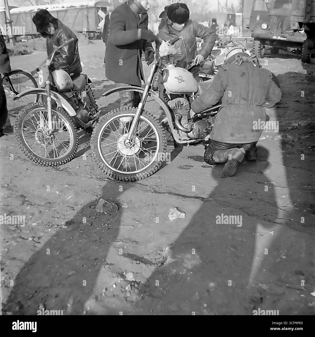 Ein Team sowjetischer Rennfahrer und Mechaniker versammelt sich, um die letzten Anpassungen an einem Motocross-Bike im Fahrerlager während eines Winterwettbewerbs in Sloviansk, der ukrainischen SSR, im Jahr 1975 vorzunehmen. Die Männer tragen schwere Mäntel und Uschanka-Hüte, um der Kälte zu trotzen, arbeiten gemeinsam an ihrer klassischen ČZ- oder Izh-Maschine. Dieses authentische Schwarzweiß-Foto fängt die düstere Atmosphäre hinter den Kulissen und die Kameradschaft des Motorsports der Basis in der UdSSR ein Stockfoto