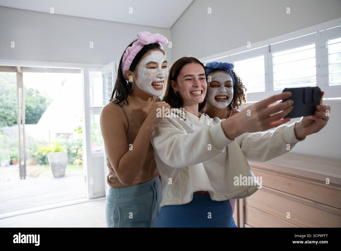Verschiedene weibliche Freunde, die zu Hause ein Smartphone für Selfie verwenden, tragen Gesichtsmasken und Stirnbänder Stockfoto