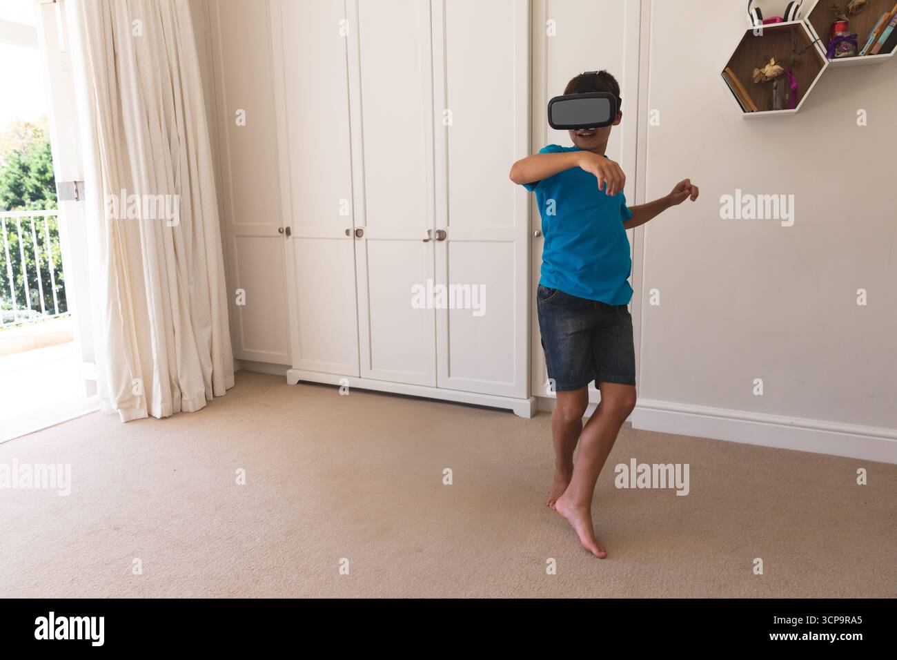 Junge im Schulalter mit blauem T-Shirt, der VR mithilfe eines Headsets im Schlafzimmer in der Nähe von Regalen und im Kopierraum erforscht Stockfoto