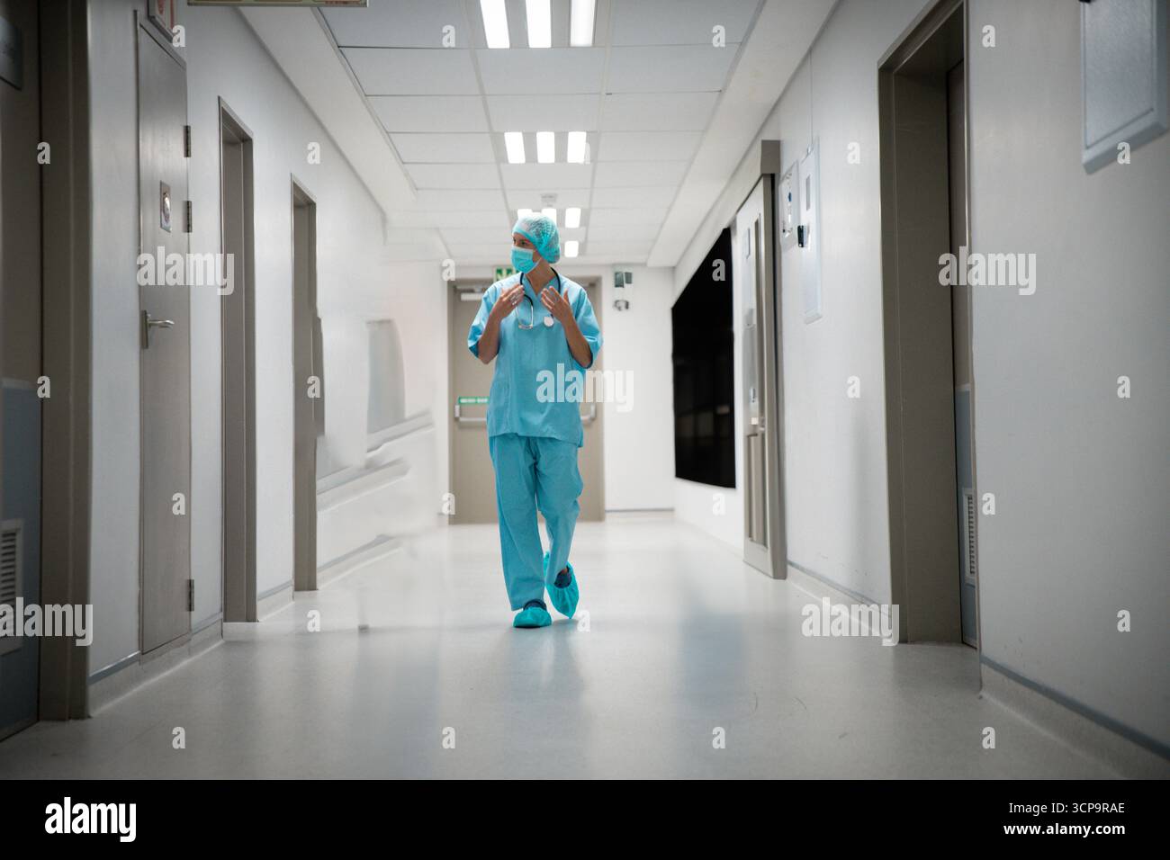 Ärztin in Peelings, die mit chirurgischer Kappe und Stethoskop die Krankenhaushalle hinuntergehen Stockfoto