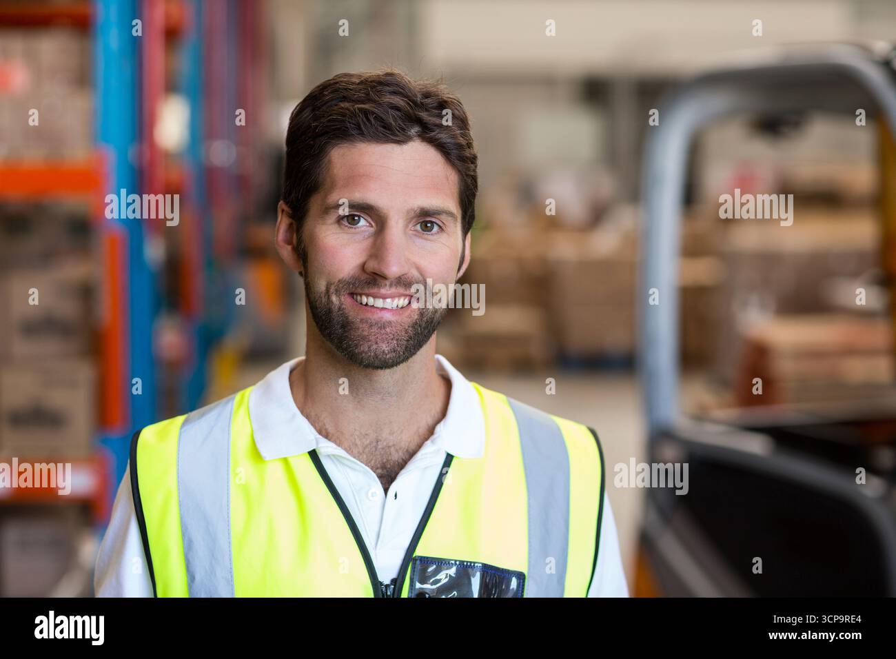 Lagerarbeiter mit gelber Weste steht im Gang mit Gabelstapler neben Regalen und Kisten Stockfoto