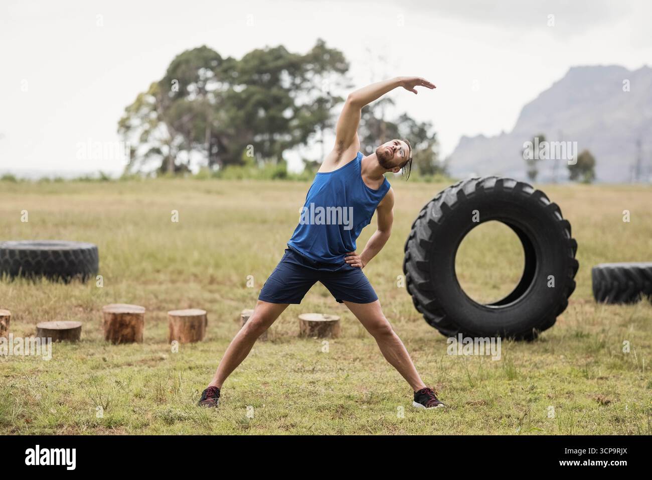 Fitnessbegeisterte Männer, die sich auf offenem Feld neben Baumstümpfen und Traktorreifen mit Laufschuhen Strecken Stockfoto