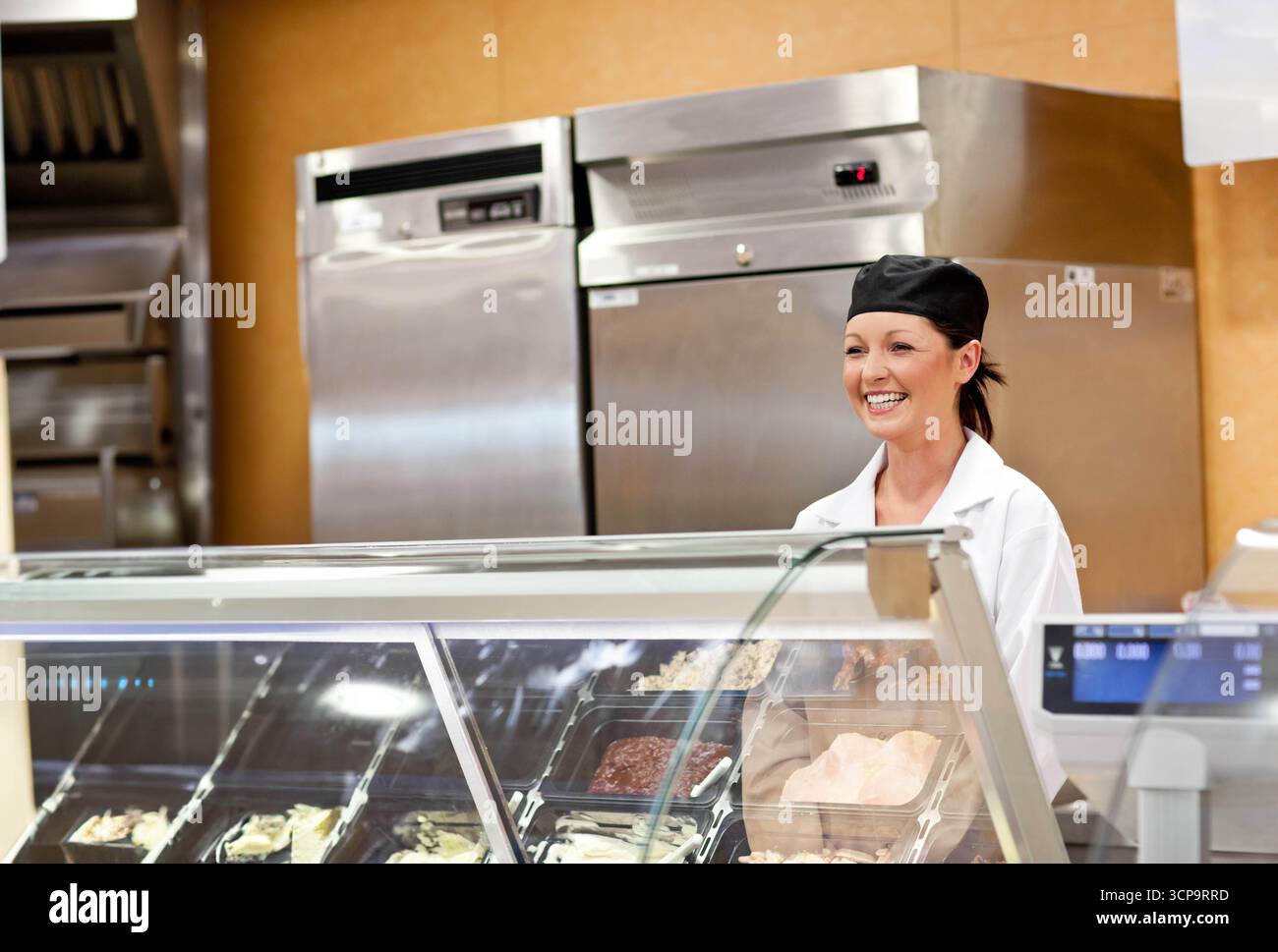 Weibliche Delikatessenarbeiterin, die hinter einem Glaskoffer mit Lebensmitteltabletts, Stahlkühlschränken und digitaler Waage arbeitet Stockfoto